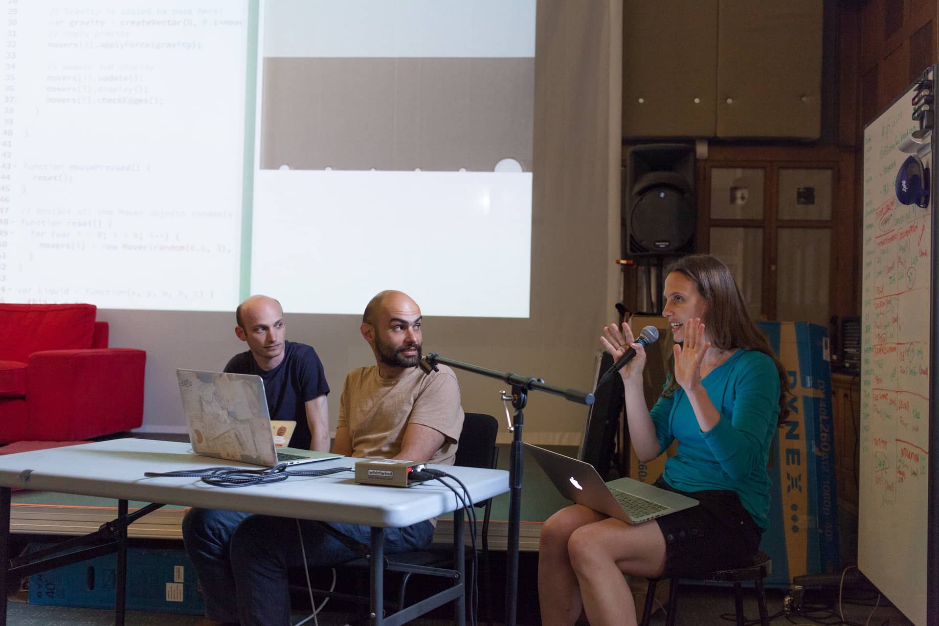 Woman expressively speaks into a microphone while two male collaborators look on"